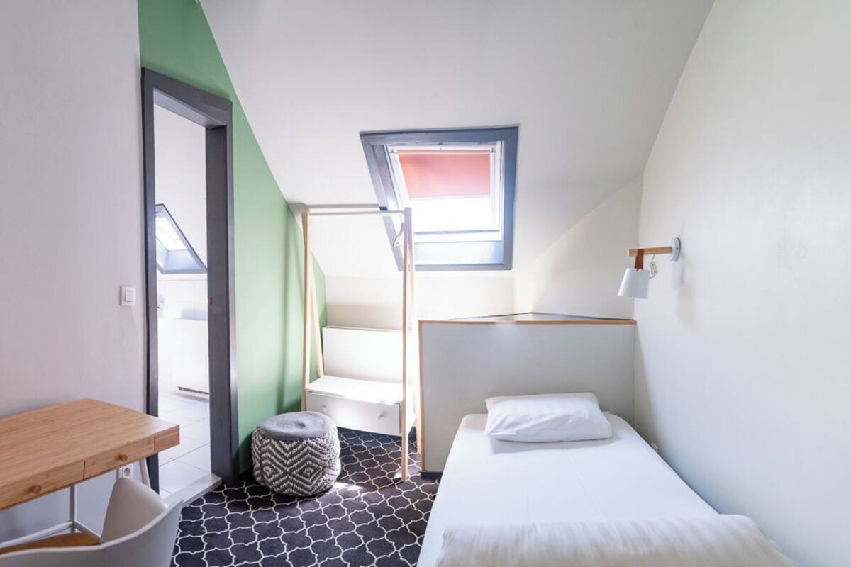 A small, modern room with a bed, a desk, and a chair. There is a window with a view of the outside, and a staircase leading to an upper level.