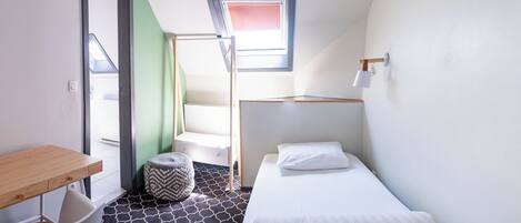 Una habitación pequeña y moderna con una cama, un escritorio y una silla. Tiene una ventana que ofrece vistas al exterior, y una escalera que sube a un nivel superior.