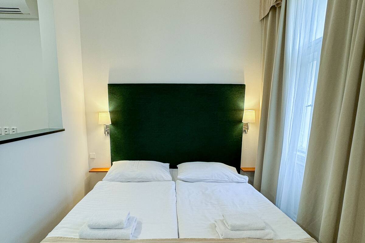 A hotel room with a bed, two bedside lamps, a green headboard, and a window with curtains.