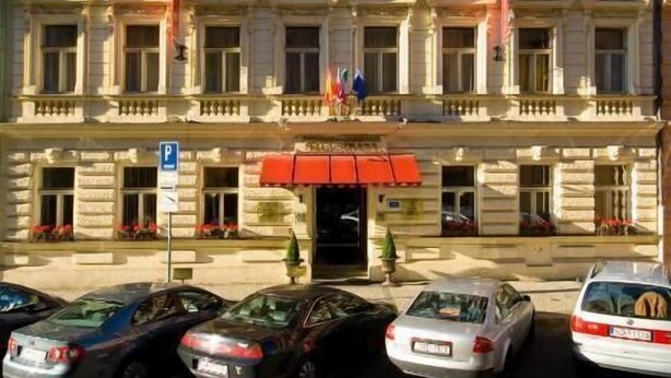 A multi-story building with a symmetrical facade, large windows, and a central entrance with a red awning. There are cars parked in front, and flags are visible on the building. - Hotel Mala Strana (Prague)