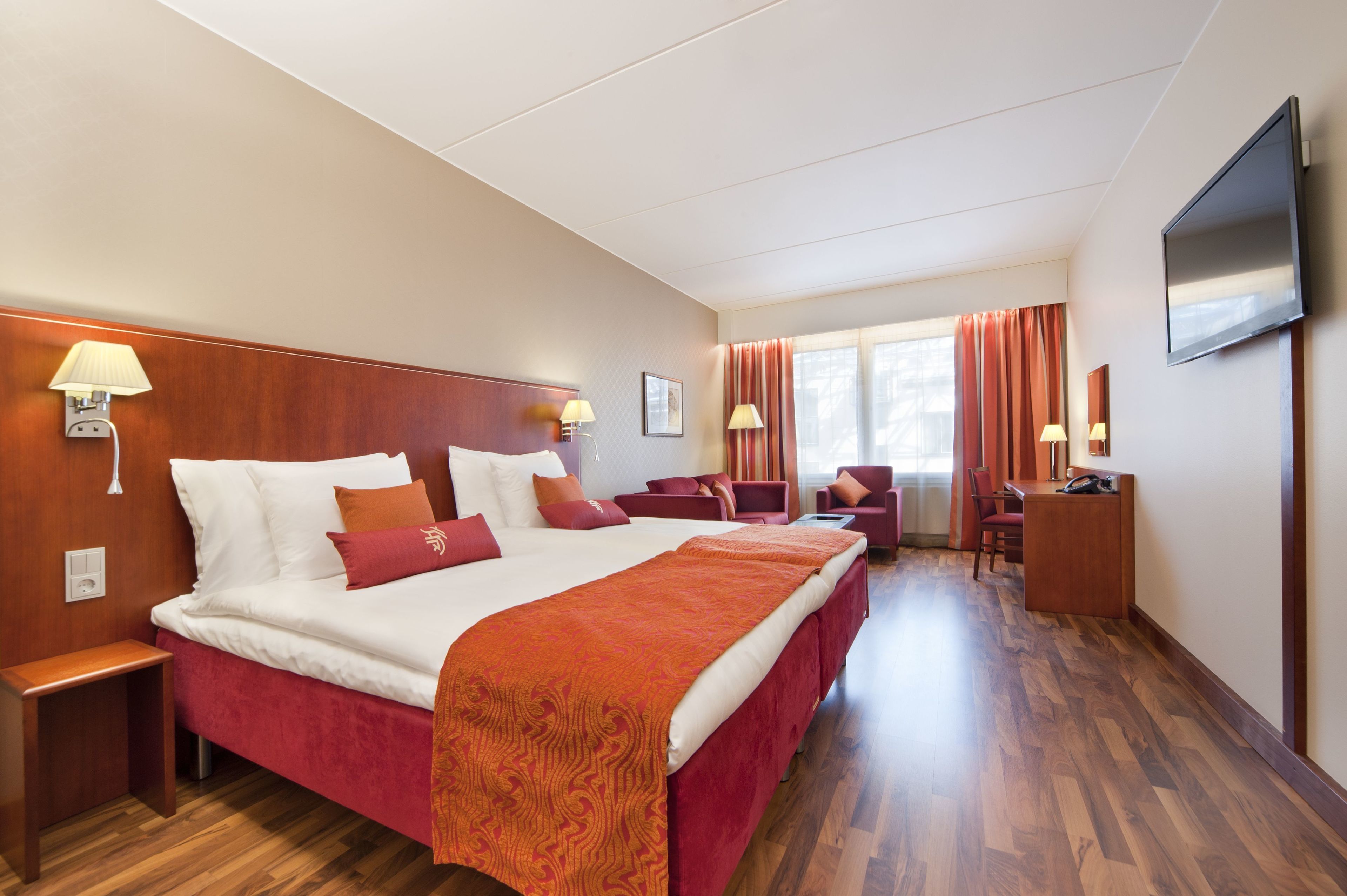 A hotel room with a large bed, a desk, two chairs, a TV, and a window with curtains.
