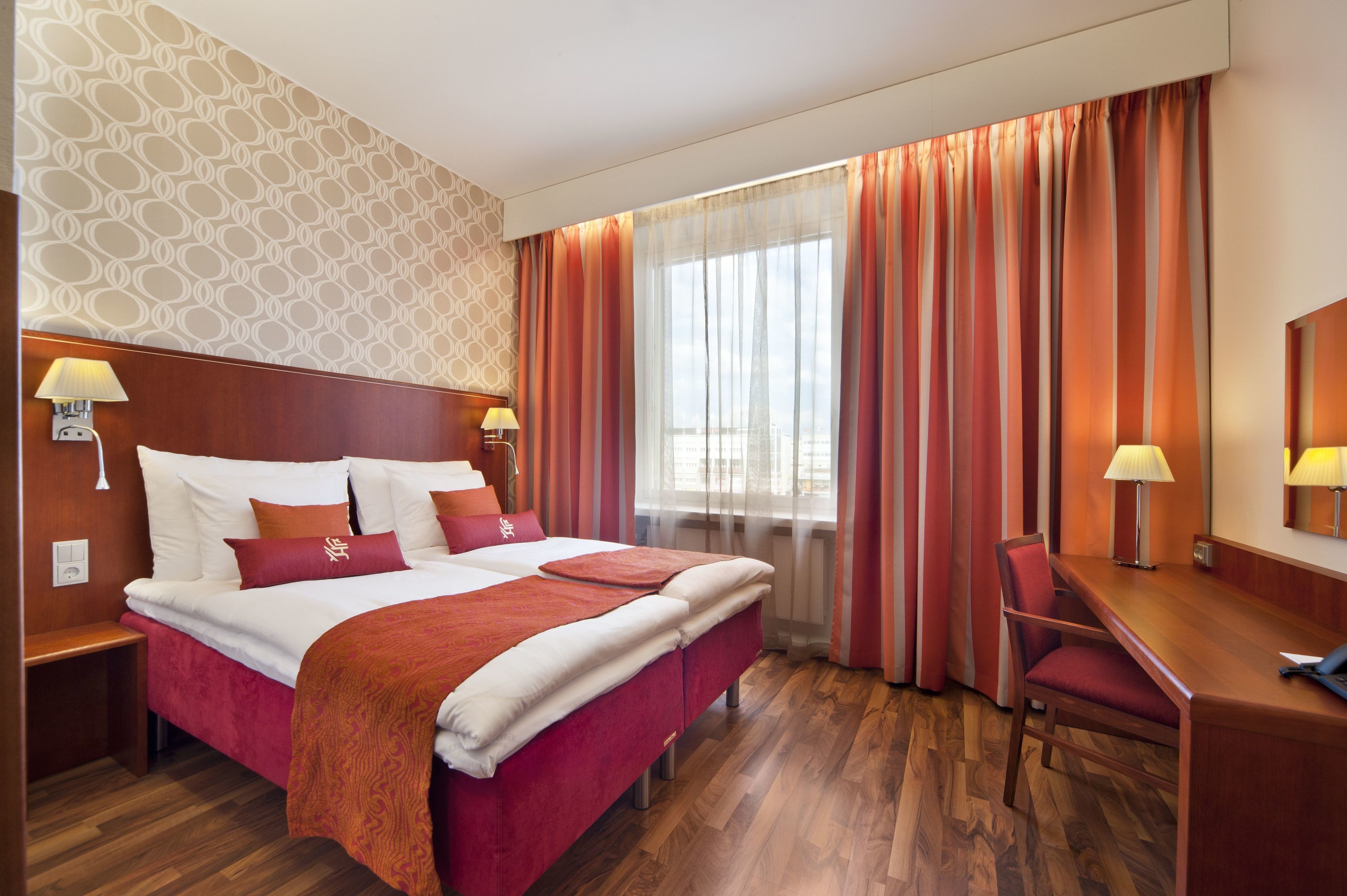 A hotel room with a large bed, a desk, and a chair. There are two lamps on the wall, and a window with curtains.