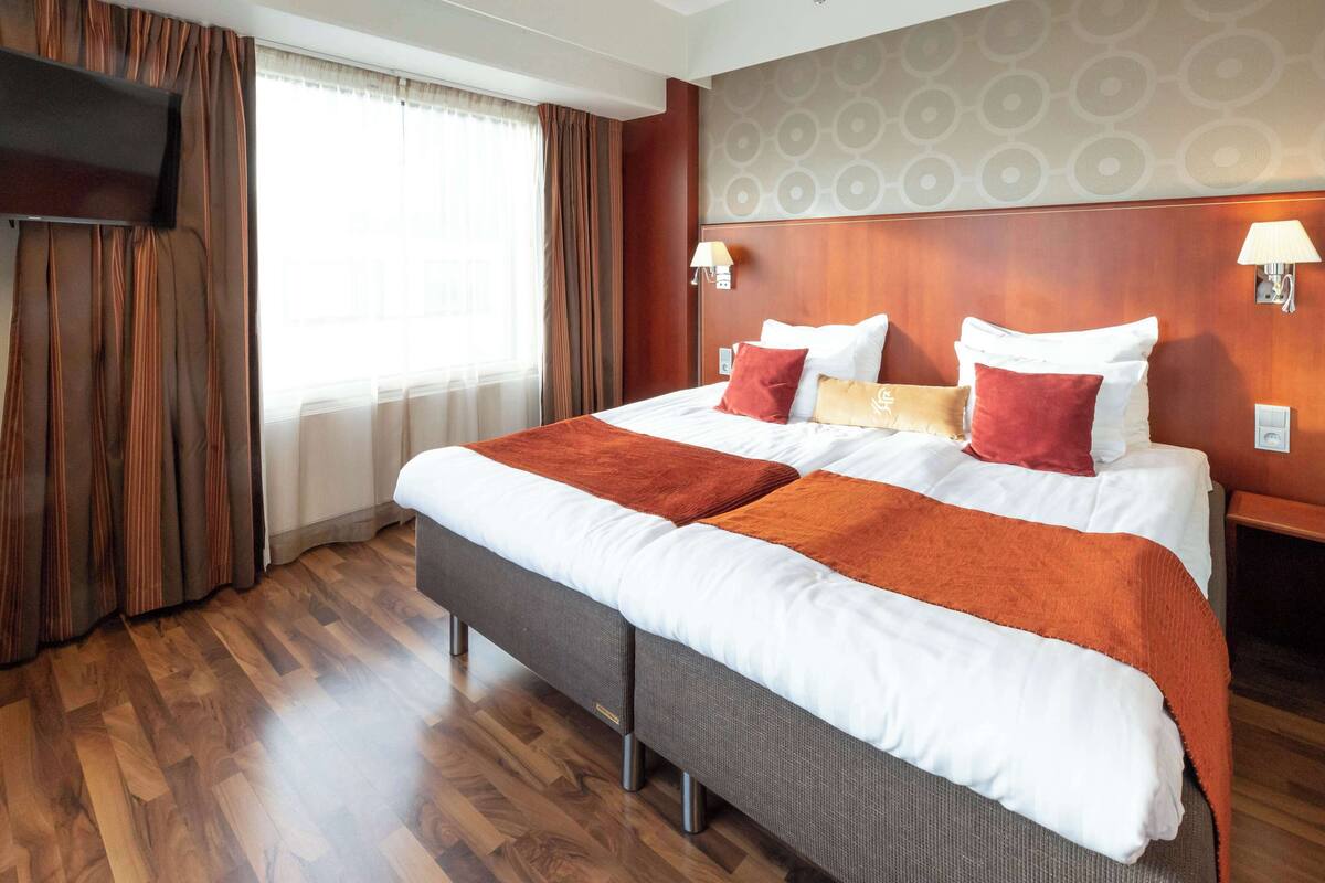Hotellihuone, jossa on suuri sänky, puulattia, seinälle asennettu televisio ja ikkuna, jossa on verhot.