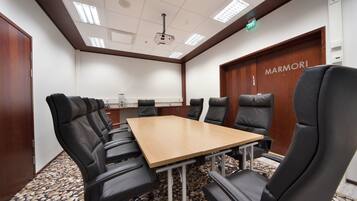 Uma sala de conferências com uma longa mesa e cadeiras de couro preto, uma porta de madeira com a placa "MARMORI" e um teto com iluminação embutida.