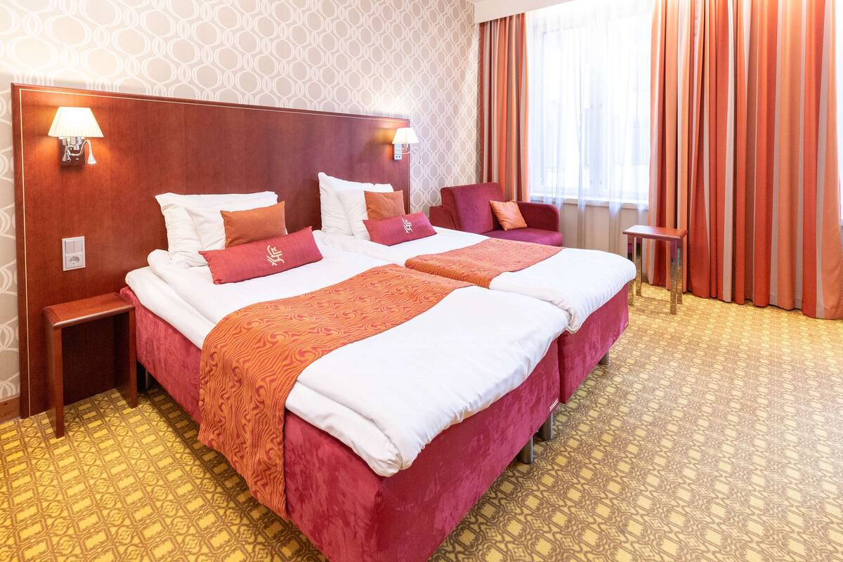 Hotellihuone, jossa on kaksi sänkyä, puinen pääty, kaksi yöpöytää, pieni sivupöytä ja kuviollinen matto.
