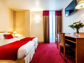 A hotel room with a bed, a desk, a chair, a TV, and a vase with flowers.