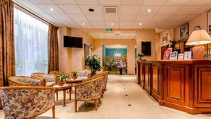 A hotel lobby with a reception desk, floral-patterned chairs, a television, and a large window with curtains.