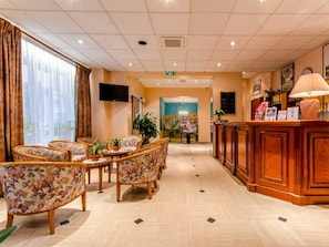 A hotel lobby with a reception desk, floral-patterned chairs, a television, and a large window with curtains.