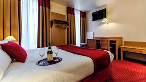 A hotel room with a large bed, a bedside table, a desk, a chair, a TV mounted on the wall, and a bottle of champagne on the bed.