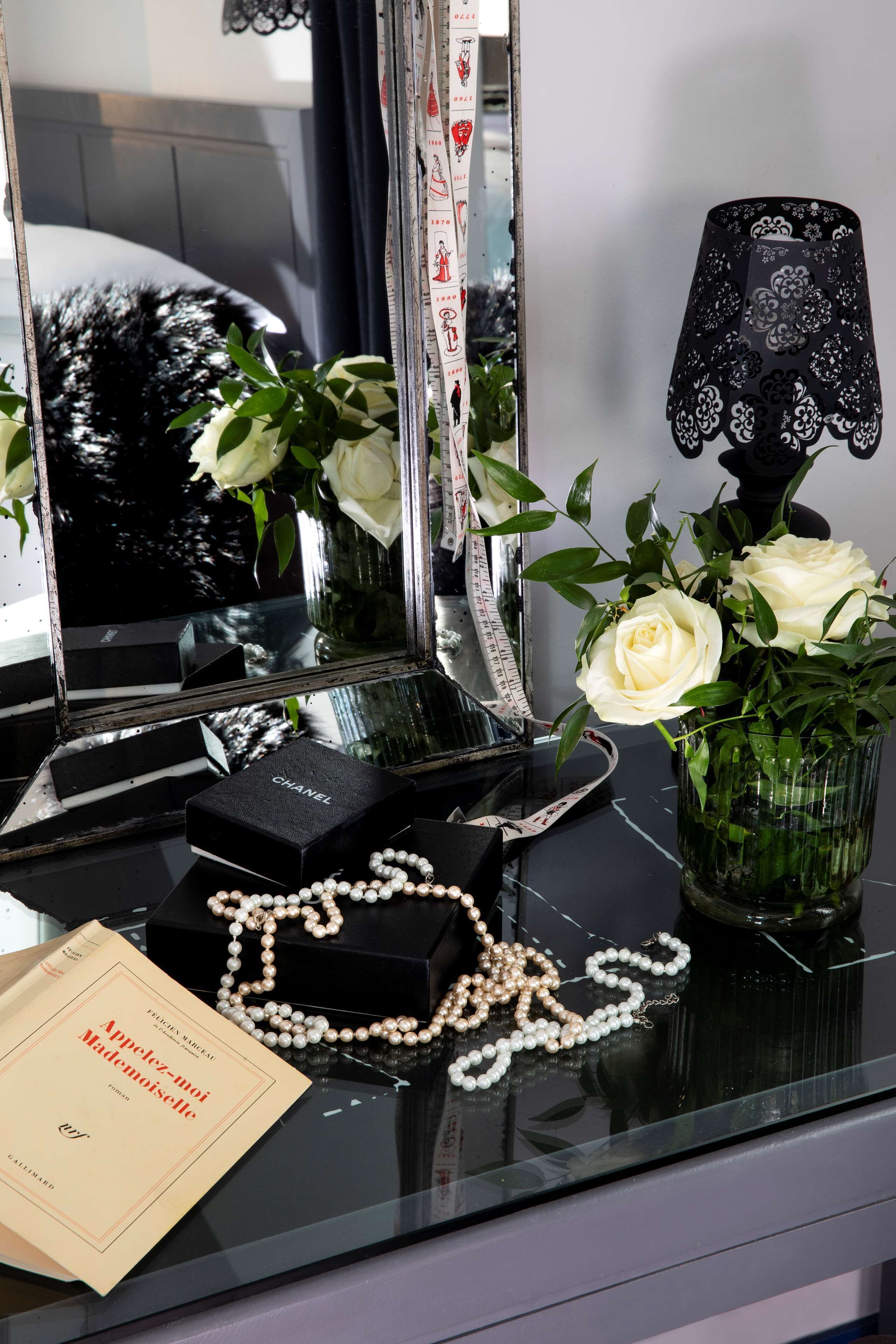 A vanity table with a mirror, a Chanel perfume box, pearl jewelry, a book titled 'Appétit-mot Mademoiselle', and a vase with white roses.