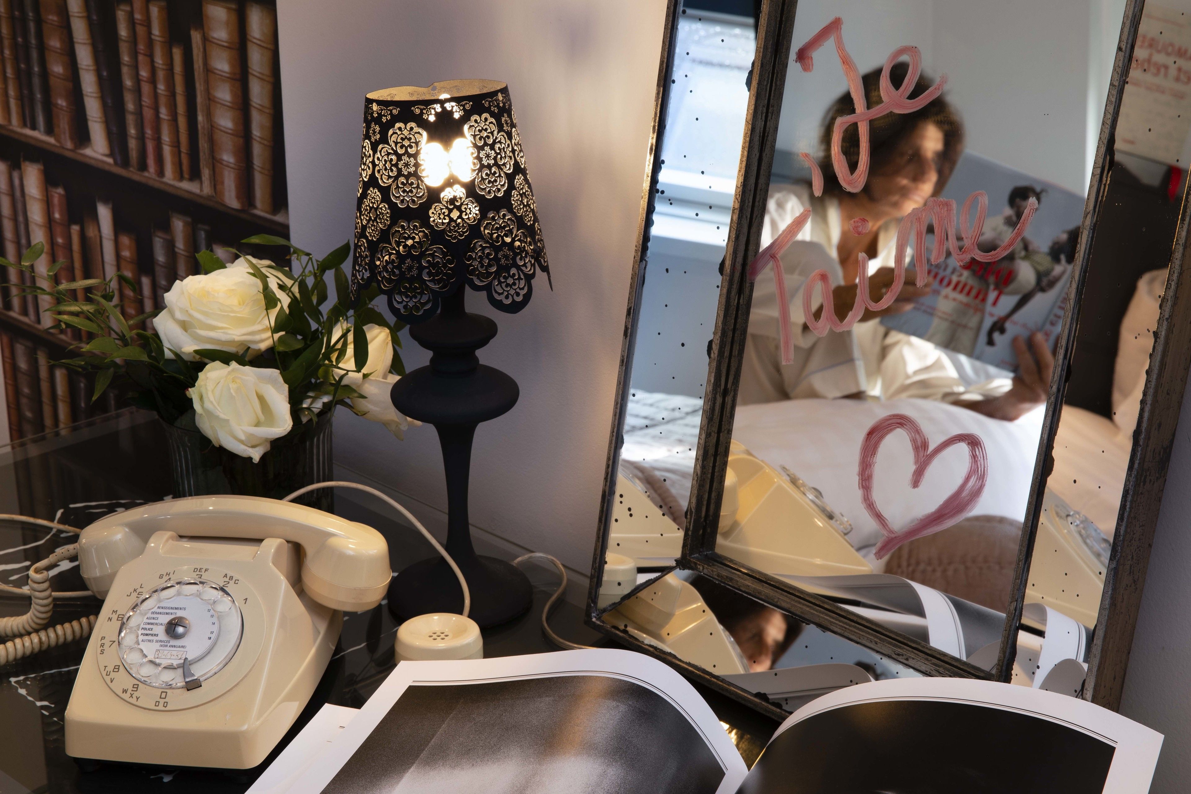 Una habitación con un teléfono rotatorio antiguo, una lámpara decorativa, un espejo con un dibujo de corazón y una estantería llena de libros.