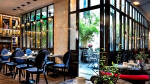 Un café moderno con suelos de madera, sillas negras y mesas con tazas y platillos. Amplias ventanas ofrecen vistas al exterior.