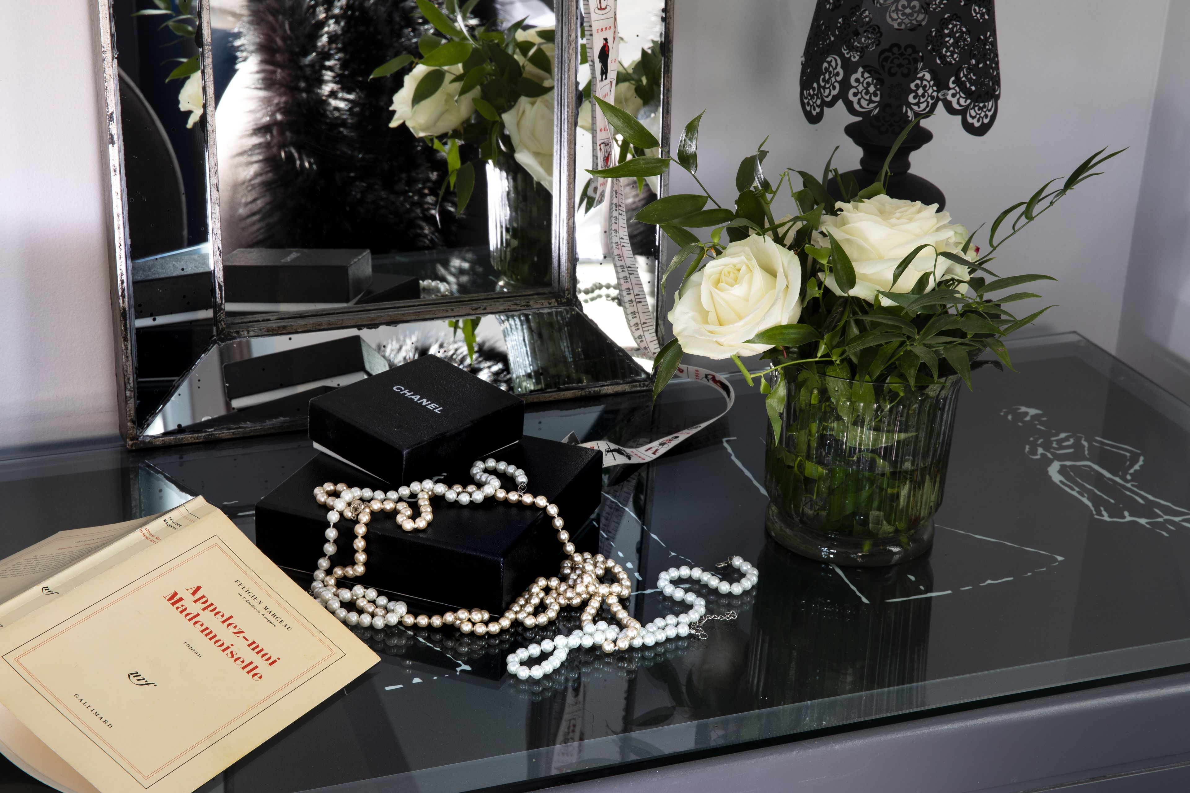 Una mesita de tocador con un espejo, un jarrón con rosas blancas, un collar de perlas, un libro titulado 'Appétit-moi' y una caja de perfume Chanel.