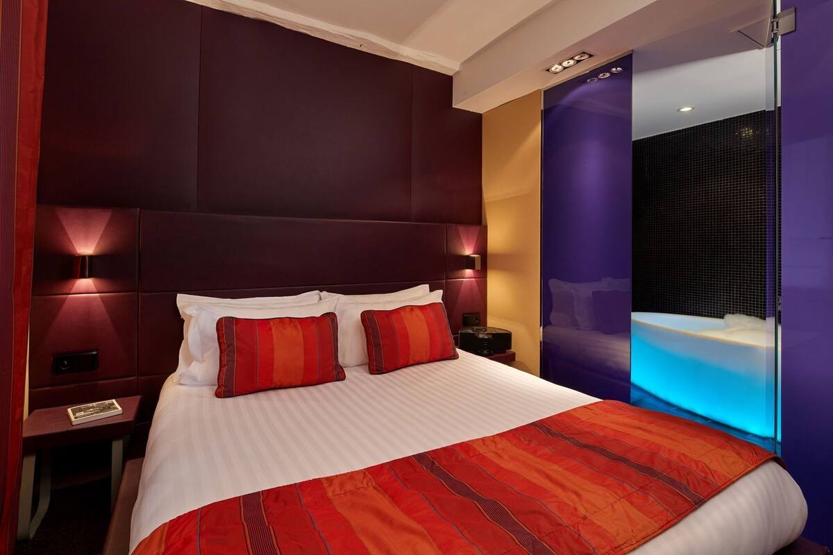 Une chambre d’hôtel avec un grand lit, deux lampes de chevet, une table de nuit et une salle de bain dotée d’une baignoire éclairée de lumière bleue.