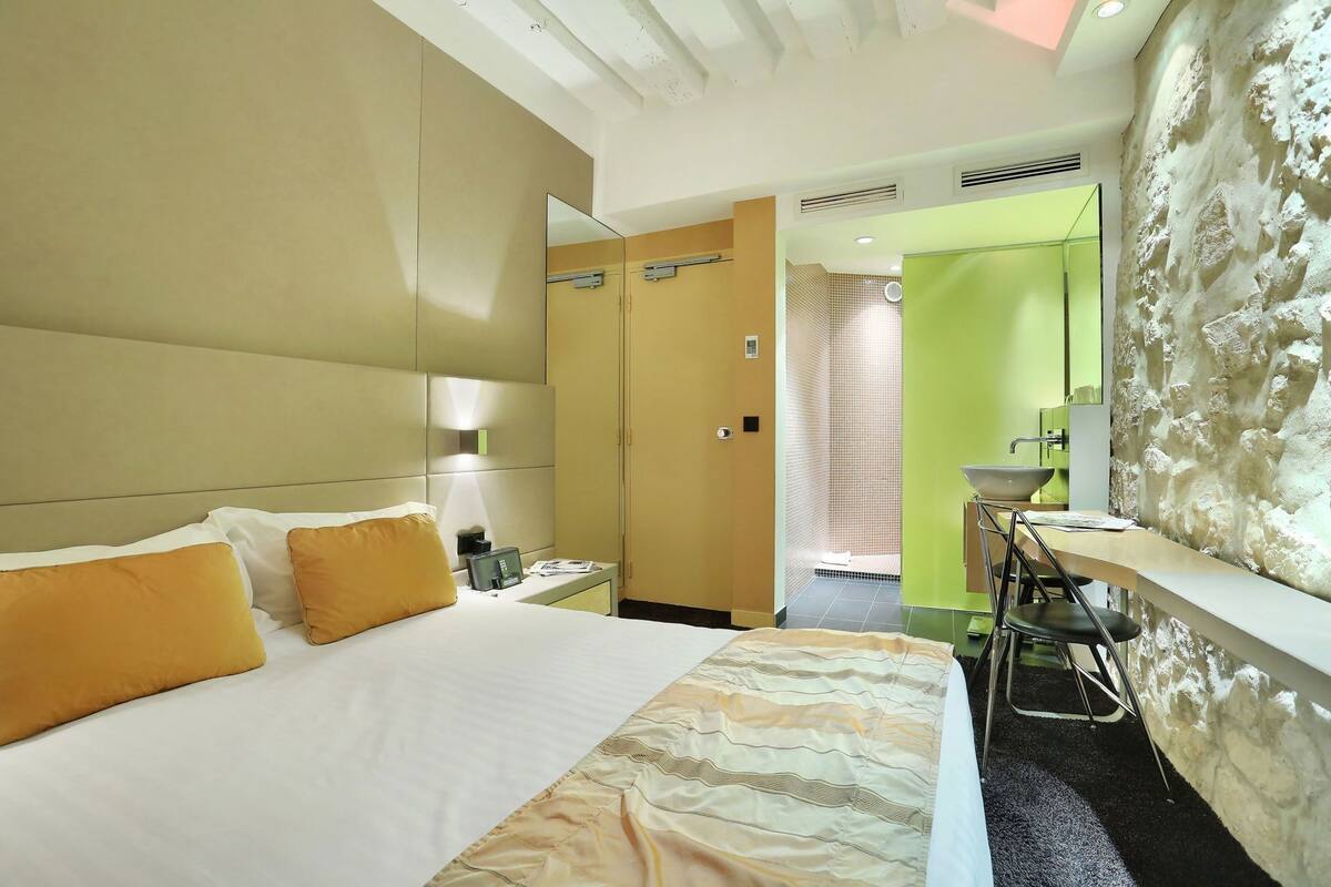 Une chambre d’hôtel comprenant un lit, une table de chevet, une lampe et une vue sur une salle de bain avec un lavabo et un miroir.