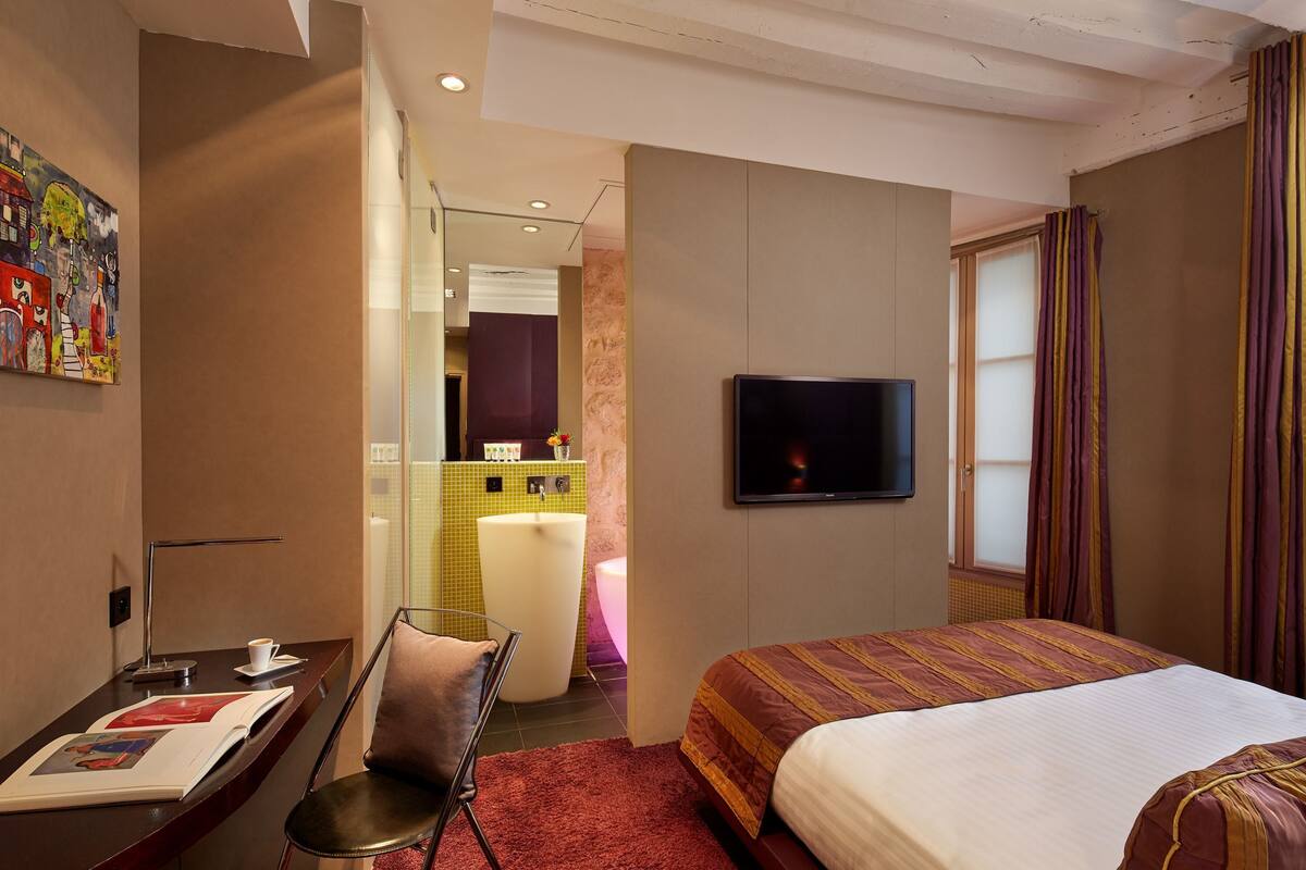 Une chambre d’hôtel comprenant un lit, une télévision, un bureau avec une chaise, une salle de bain avec un lavabo et un miroir, et un tableau accroché au mur.