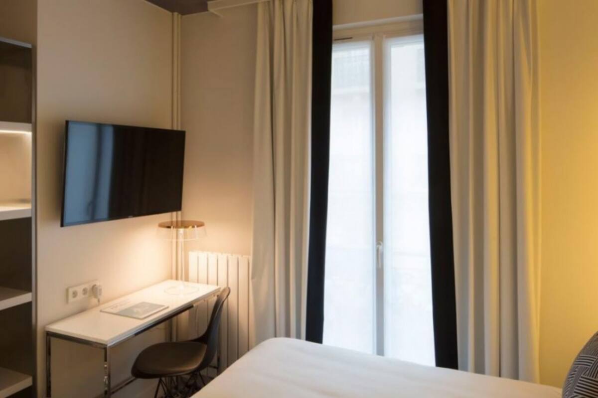 Ein Hotelzimmer mit einem Bett, einem Schreibtisch mit Stuhl, einem an der Wand montierten Fernseher und einem Fenster mit Vorhängen.