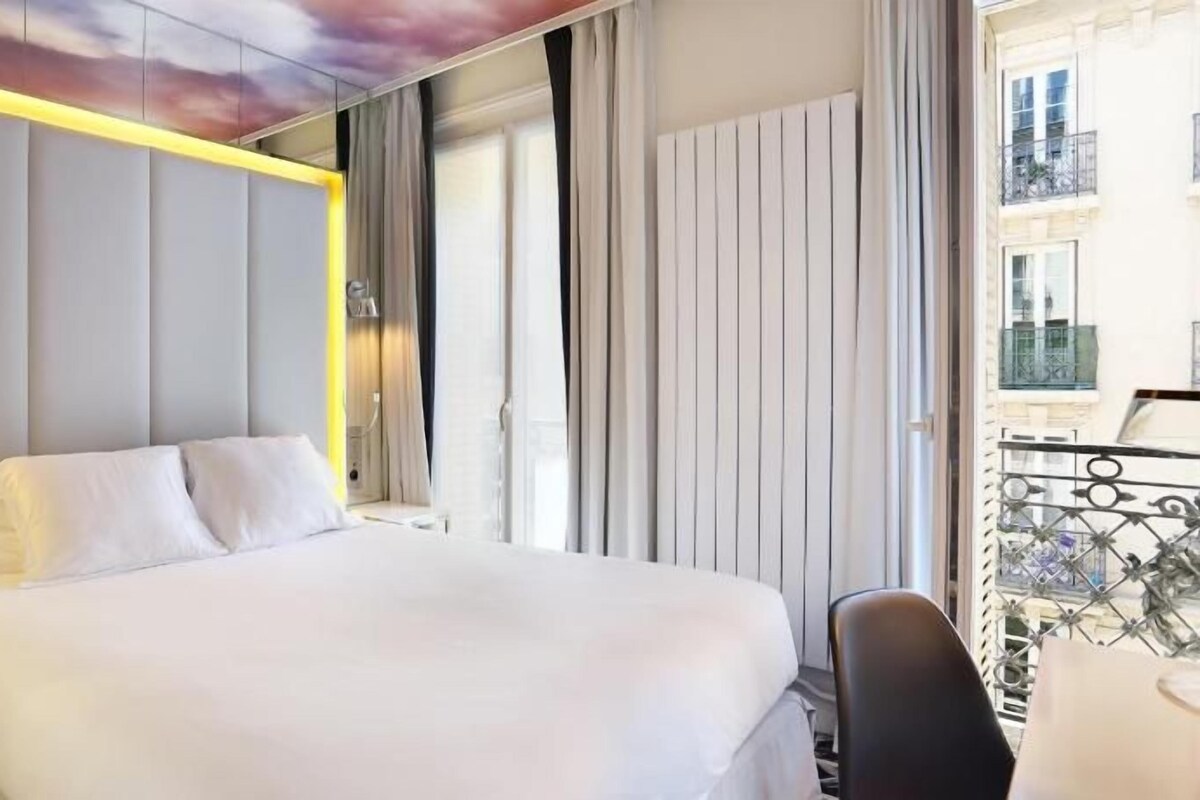 A hotel room with a large bed, a bedside table, a chair, and a view of the city.