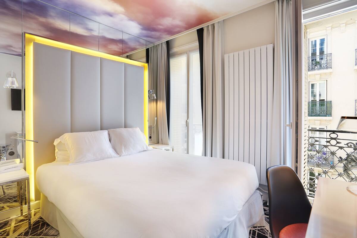 Ein Hotelzimmer mit einem großen Bett, einem Schreibtisch mit Lampe und Blick auf die Stadt durch das Fenster.