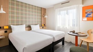 Habitación de hotel con dos camas, una mesita pequeña con una silla, una ventana con cortinas ligeras y una pared con estampado escocés.
