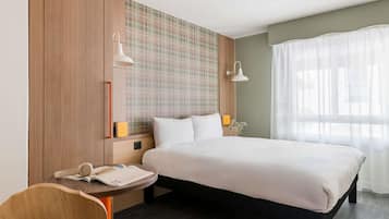 Une chambre d’hôtel comprenant un lit, des tables de chevet, un bureau, une chaise, une lampe fixée au mur et une fenêtre avec des rideaux.