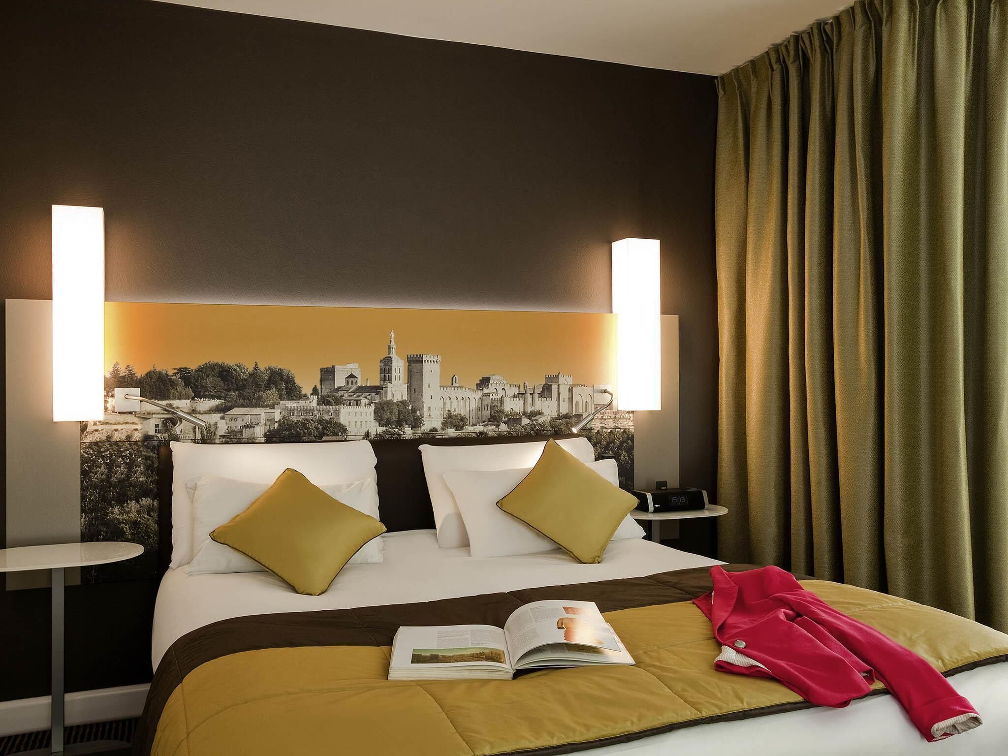 Une chambre d’hôtel avec un lit, deux tables de chevet, une œuvre d’art représentant un paysage urbain au niveau de la tête de lit, et une fenêtre avec des rideaux.