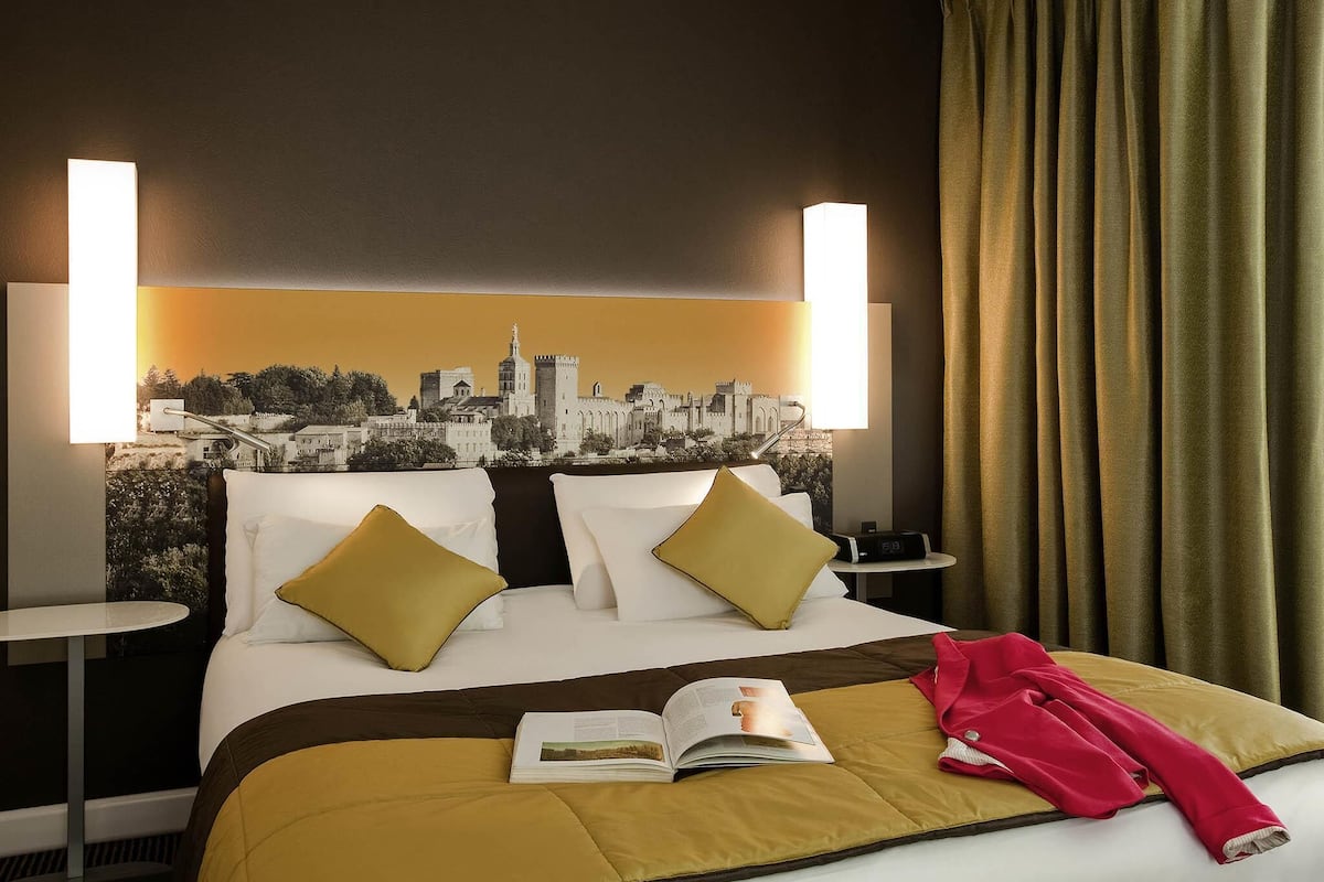 A hotel room with a bed, two bedside tables, a cityscape artwork on the headboard, and a window with curtains.