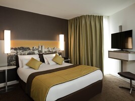 A hotel room with a bed, two bedside tables, a TV, a cityscape mural, and a window with curtains.