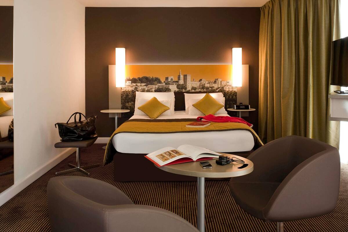 Habitación de hotel con cama, dos sillas, una mesita, una mesita de noche y un cuadro de arte con vista a la ciudad.