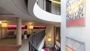 Um saguão de hotel moderno com uma escada em curva, uma área de estar com sofá e poltronas, e um grande quadro abstrato na parede.