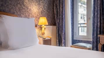 Una habitación de hotel con cama, mesita de noche con lámpara, ventana con cortinas y vista a edificios.