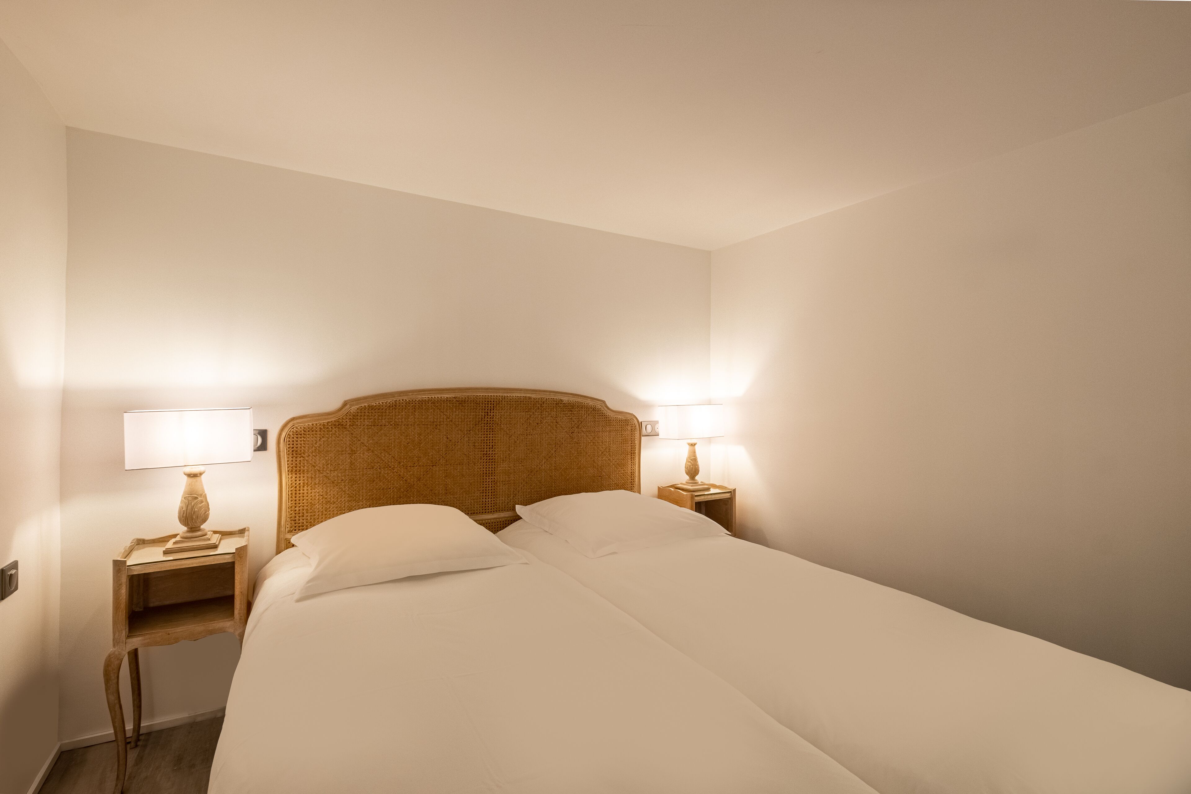 A neatly made bed with white linens in a room with white walls and a wooden headboard. There are two bedside tables with lamps on either side of the bed.