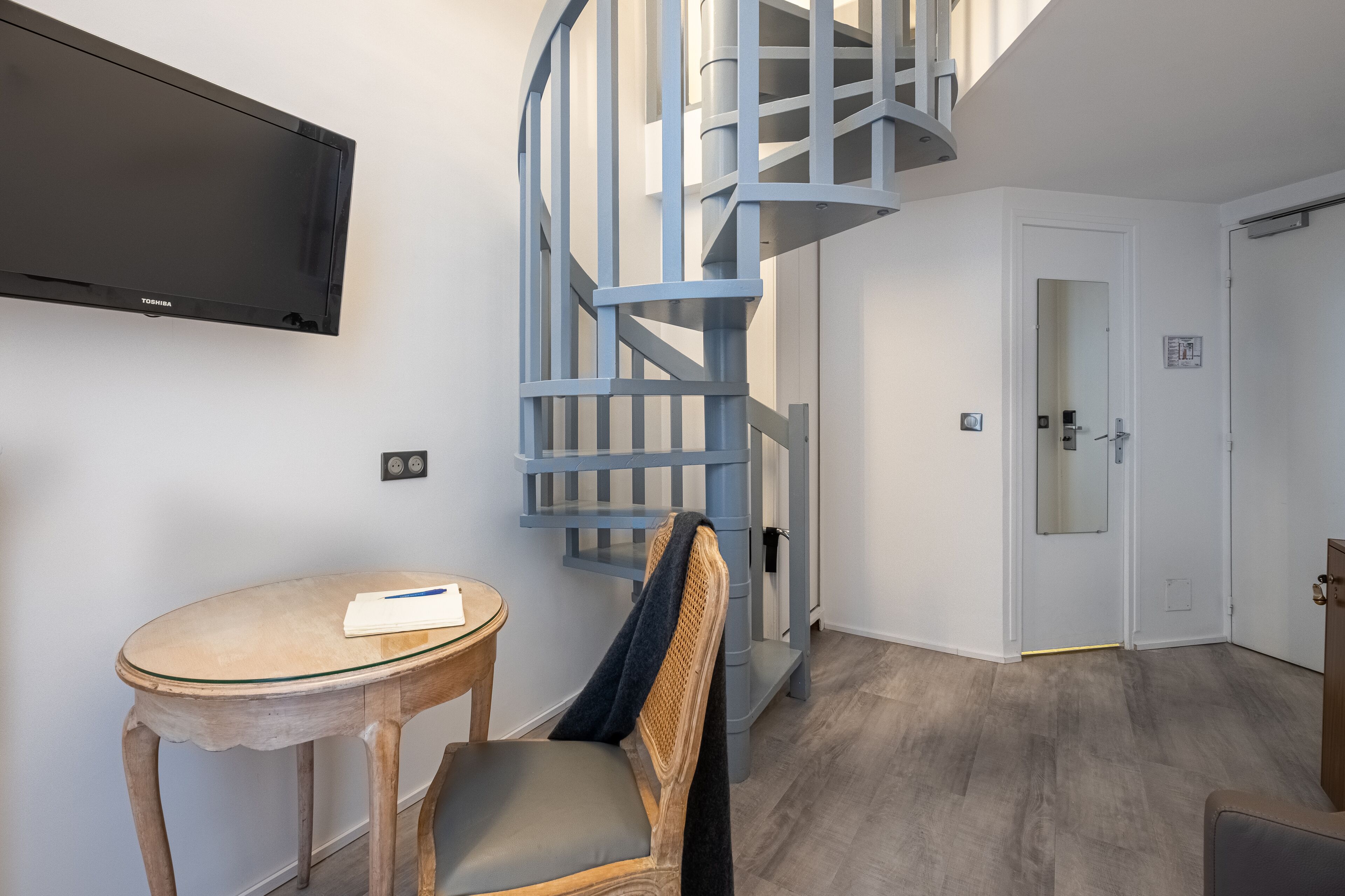 A modern room with a spiral staircase, a flat-screen TV mounted on the wall, a round table with a chair, and a door leading to another room.