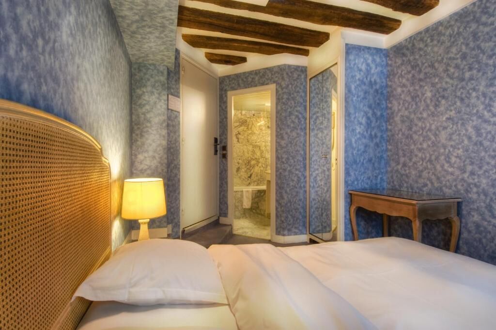 Habitación de hotel con cama, mesita de noche, lámpara, cabecero de madera, espejo y puerta que da a un baño.