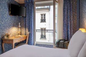 A hotel room with a bed, a desk with a lamp, a TV mounted on the wall, and a view of a building through the window.