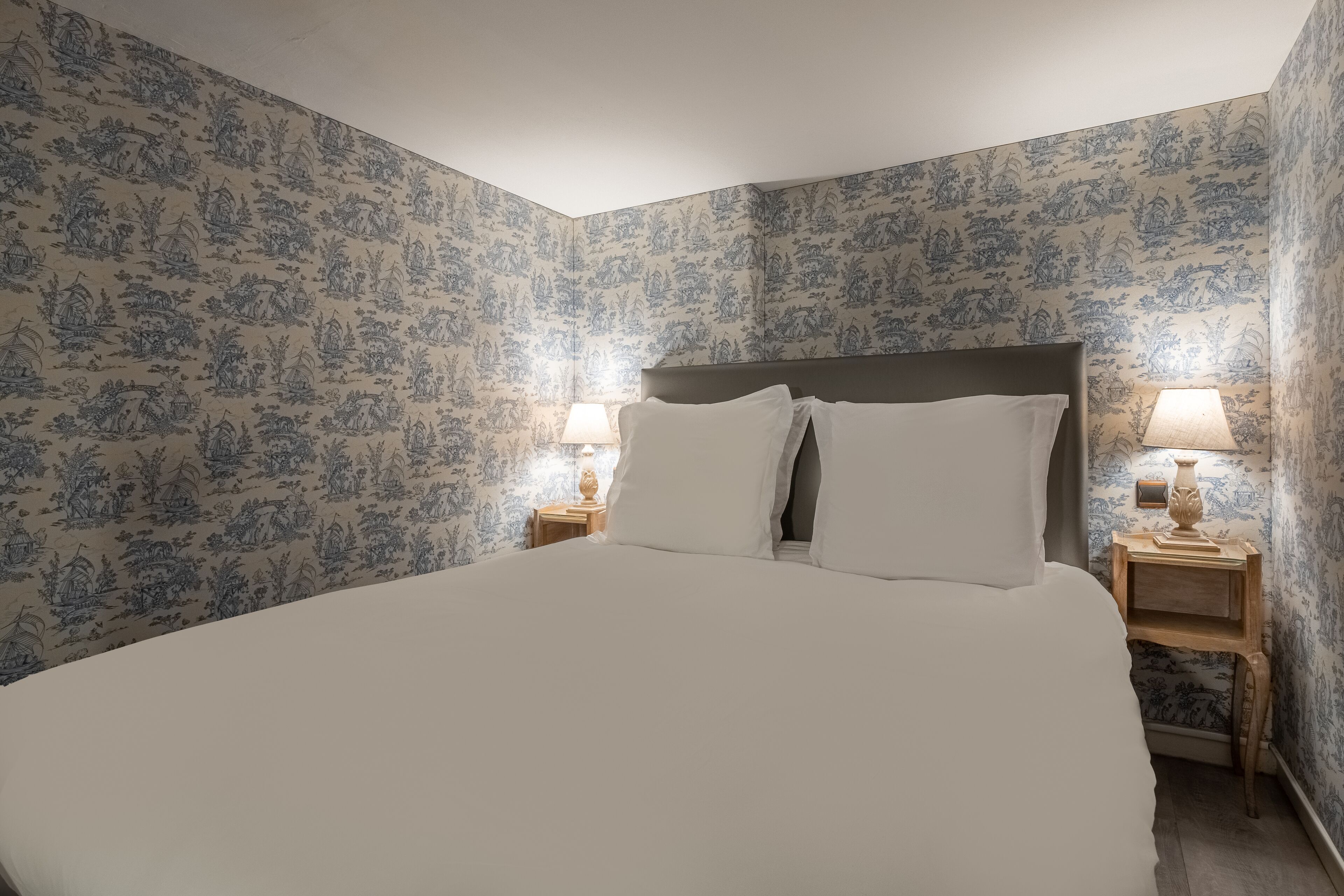 Una cama bien hecha con ropa blanca en una habitación con papel tapiz floral. Hay dos mesitas de noche con lámparas.