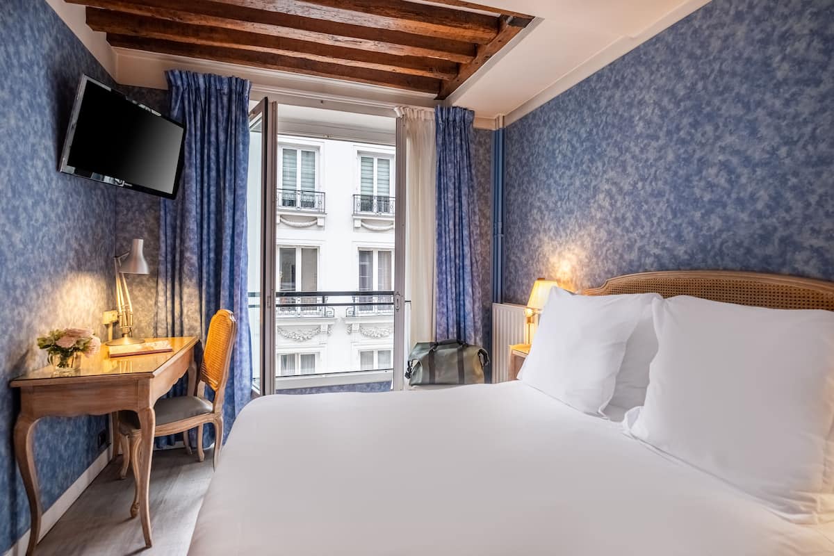 A hotel room with a bed, a desk with a lamp, a chair, a TV, and a view of the outside balcony.