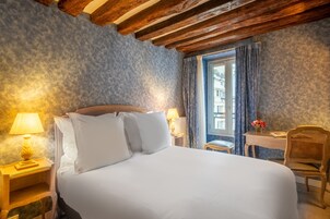 Ein Hotelzimmer mit einem großen Bett, Holzbalkendecke, einem Fenster mit Vorhängen, einem kleinen Tisch mit Stuhl und einer Lampe auf einem Nachttisch.