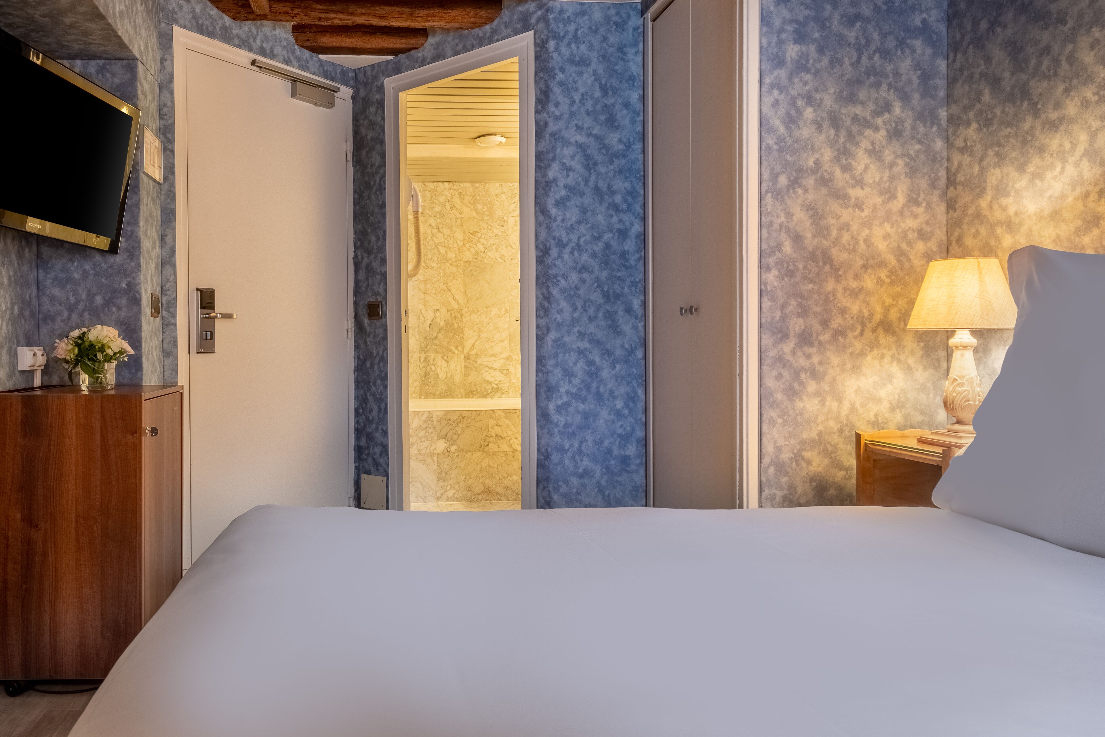 Una camera da letto con un letto, una cassettiera in legno, una TV montata a parete e un bagno visibile attraverso una porta aperta.