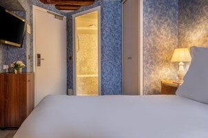 A bedroom with a bed, a wooden dresser, a TV mounted on the wall, and a bathroom visible through an open door.