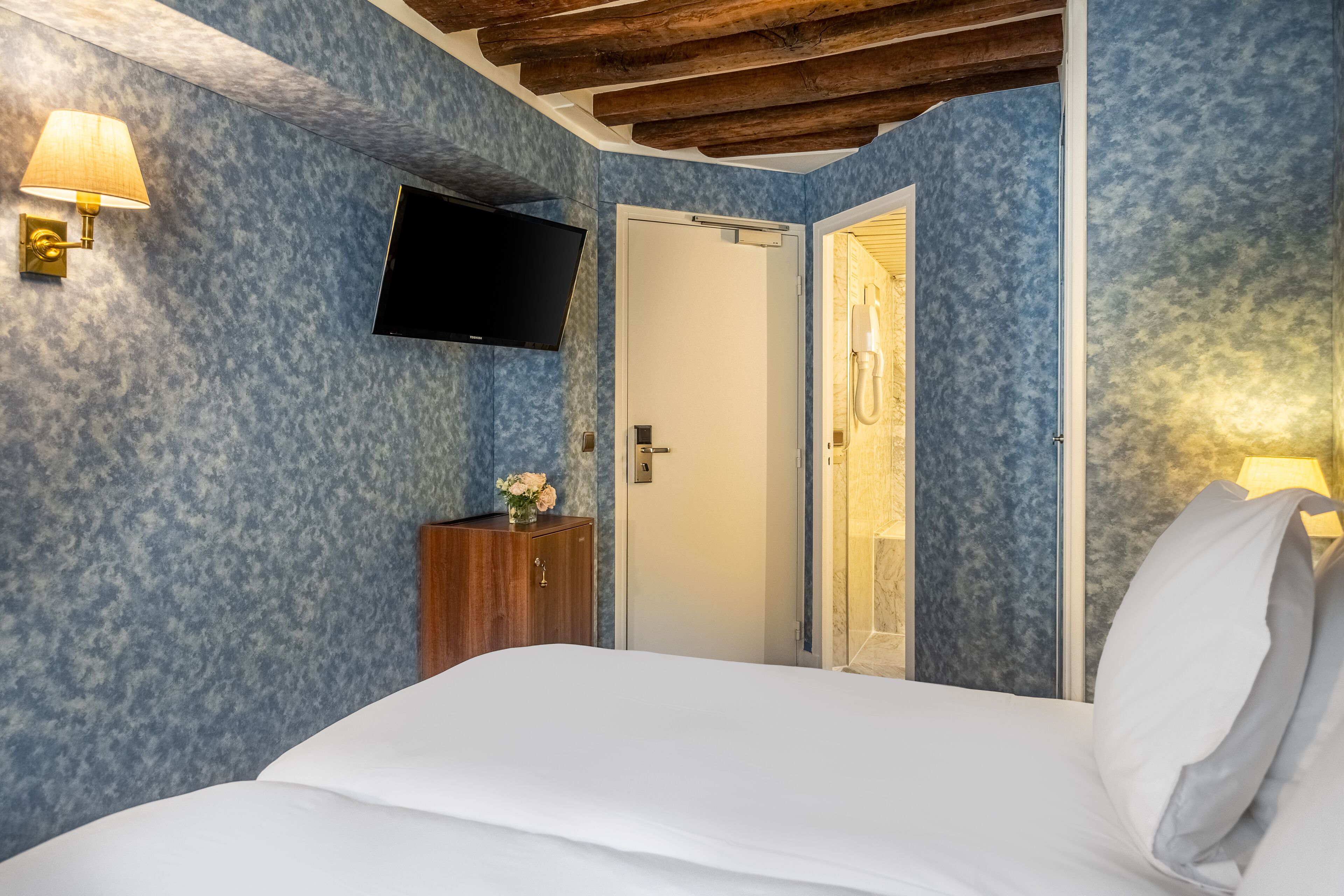 A bedroom with a bed, a TV mounted on the wall, a wooden cabinet, and a door leading to a bathroom.