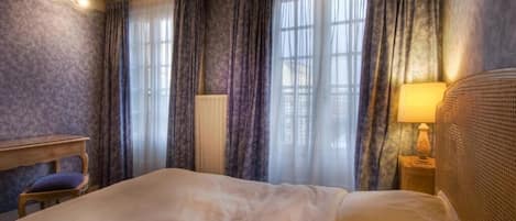 Quarto de hotel com uma cama grande, mesa de cabeceira de madeira com abajur, uma cadeira e uma janela com cortinas.