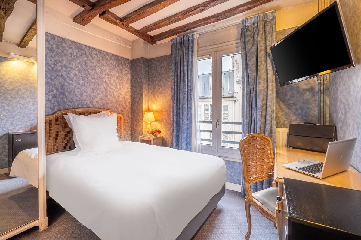A hotel room with a bed, a desk with a laptop, a chair, a TV, and a window with curtains.