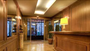 Un vestíbulo de hotel con paneles de madera, un mostrador de recepción, una planta en maceta y una puerta de cristal que da a la entrada.