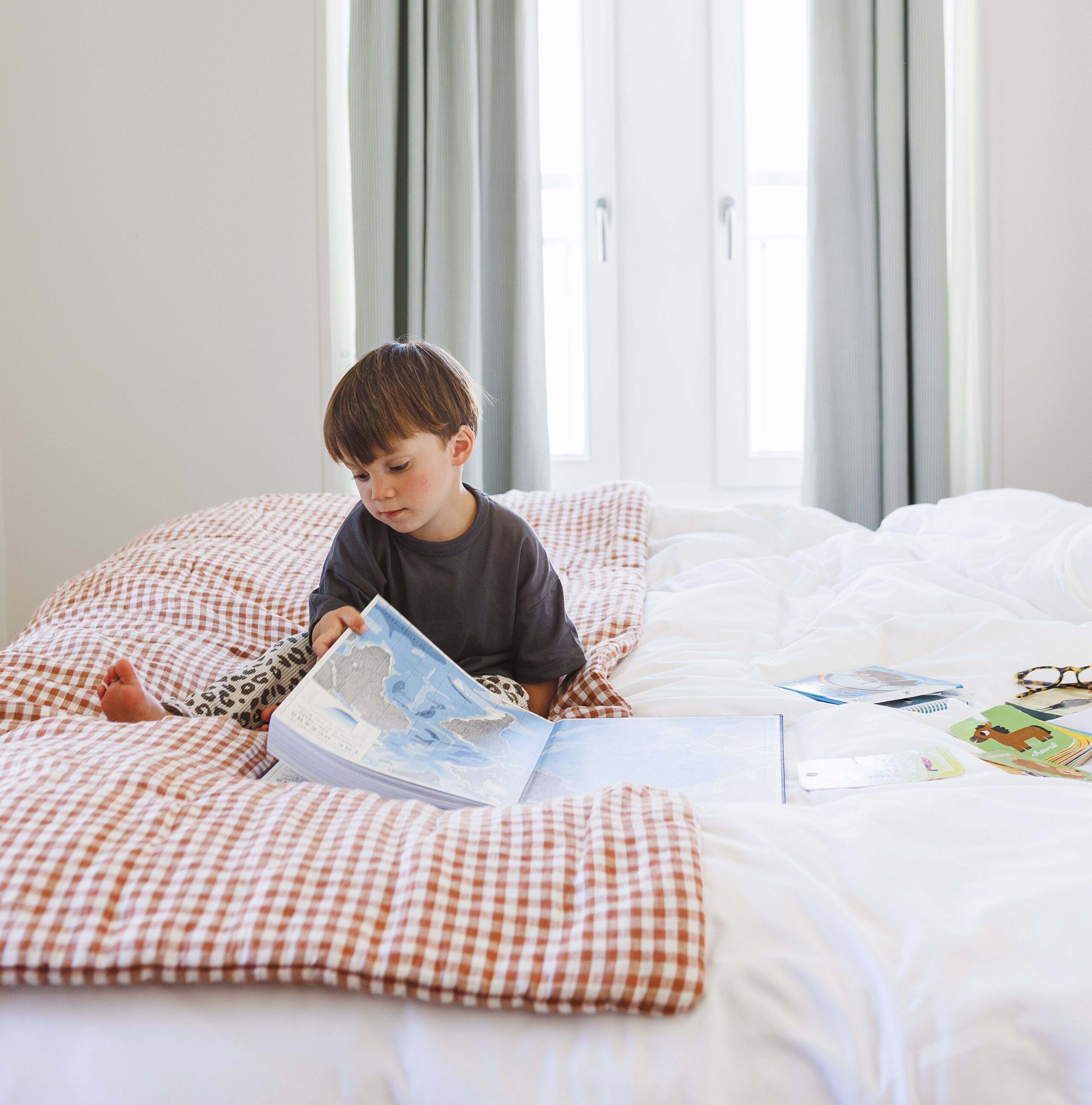 A child sitting on a bed with a book, in a room with checkered bedding and curtains.