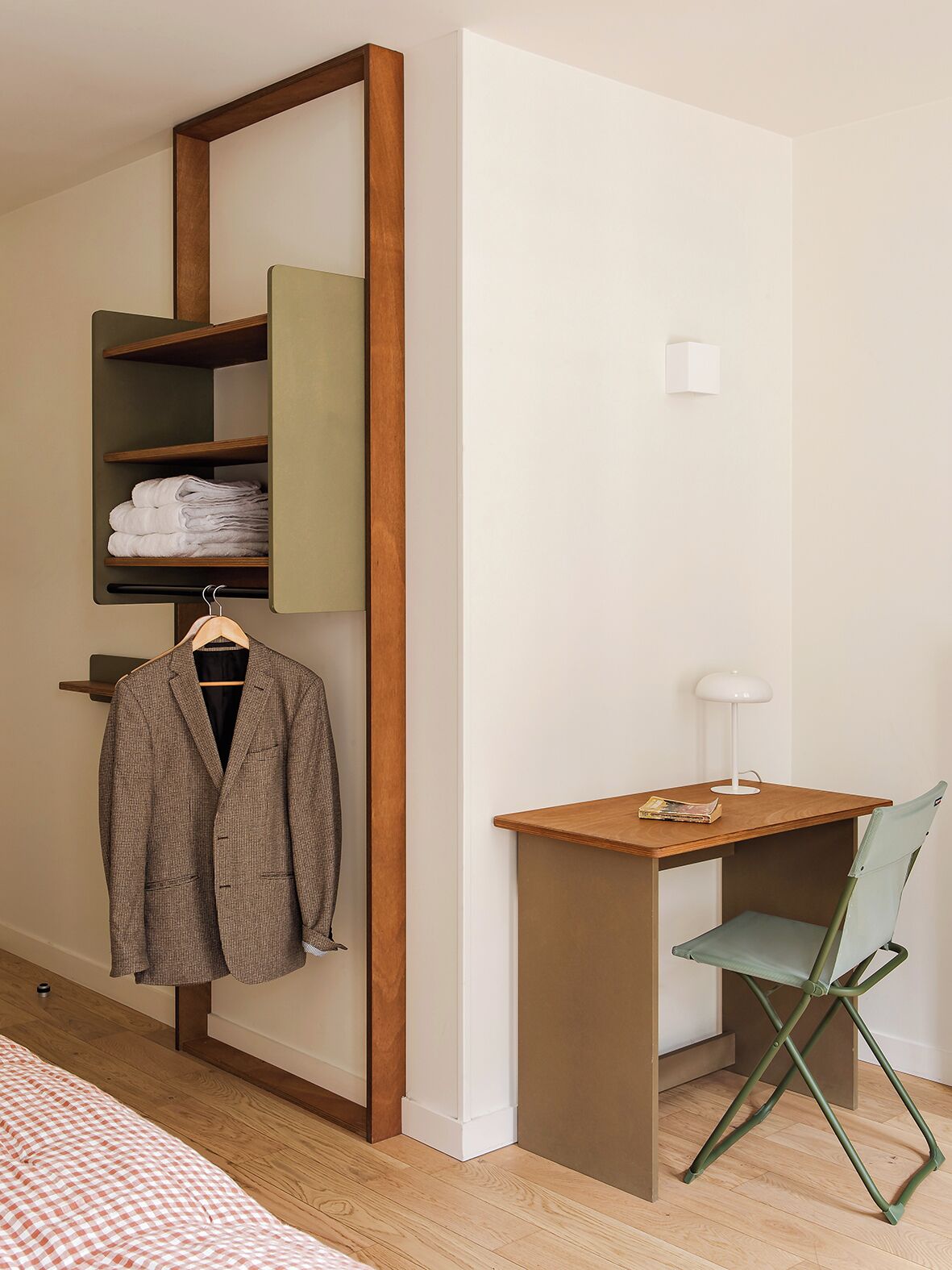A compact room with a wooden desk, a green folding chair, a hanging coat, and a shelf with folded towels.