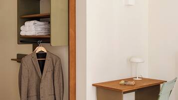 A compact room with a wooden desk, a green folding chair, a hanging coat, and a shelf with folded towels.