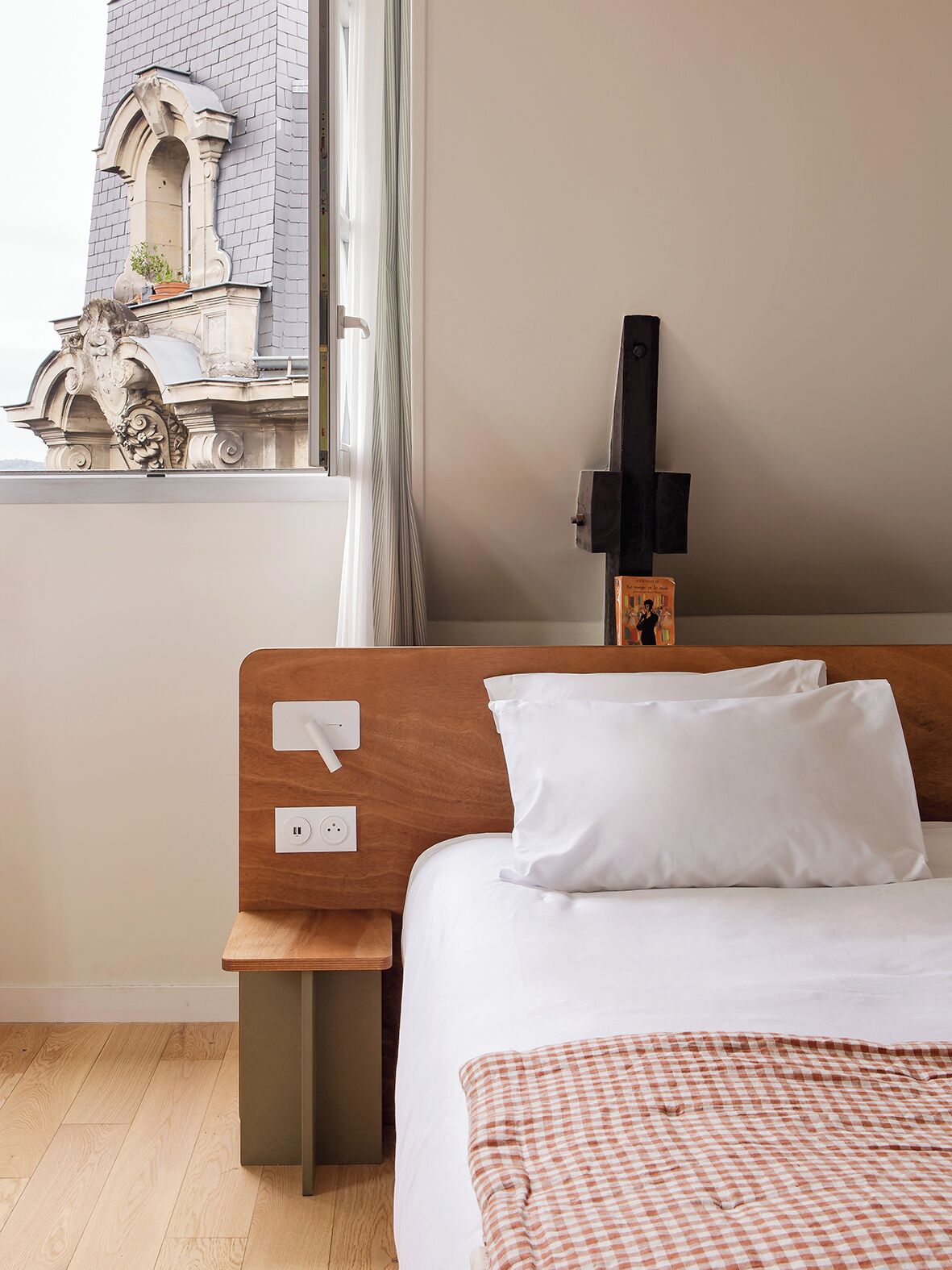 A hotel room with a wooden headboard, a white pillow, a checkered blanket, and a view of a building with a window and a curtain.