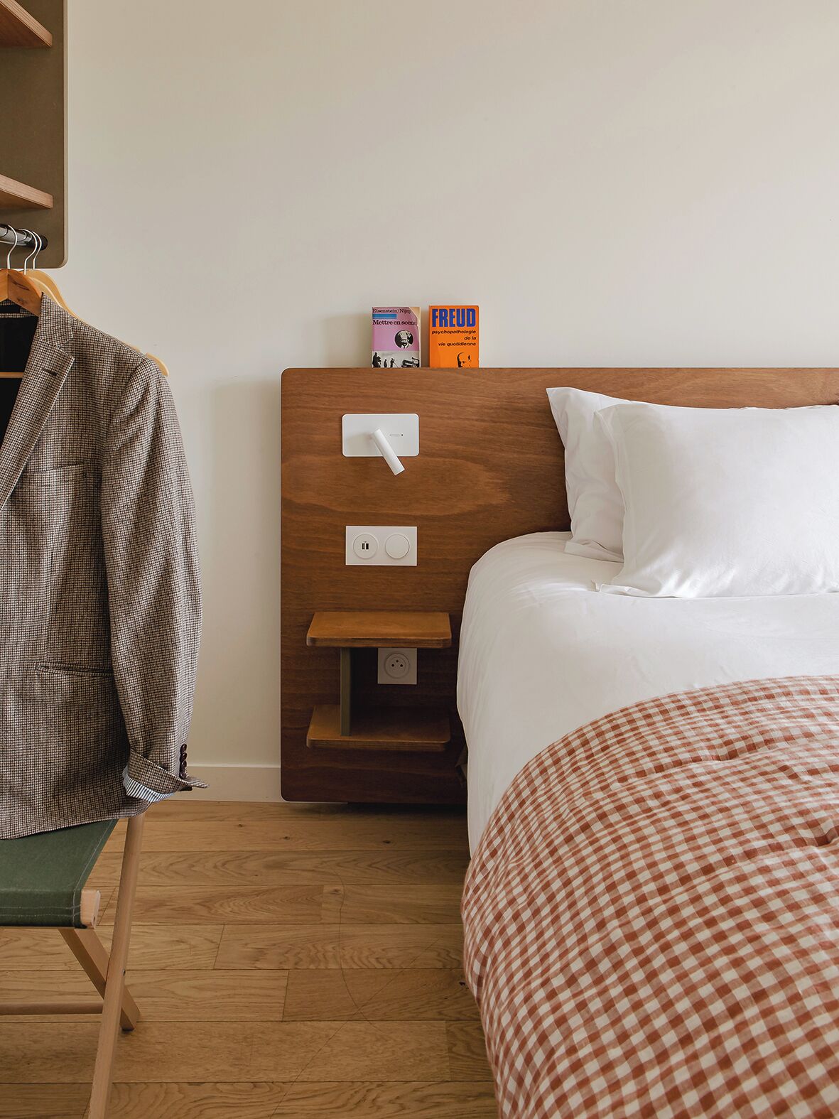 Una cama bien hecha con ropa de cama a cuadros blancos y rojos, un cabecero de madera y una mesita de noche con un libro y un control remoto.