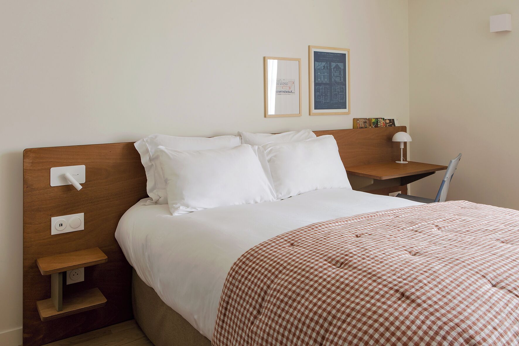A neatly made bed with white linens and a checkered blanket, a wooden nightstand with a lamp, and a small table with a chair.